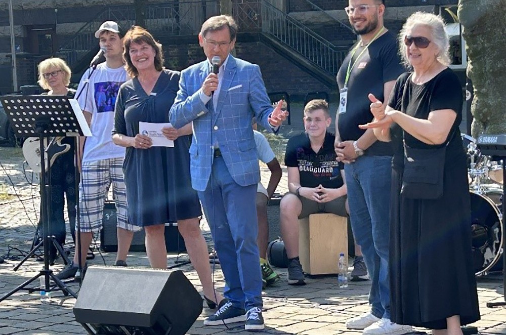 Das zweite Foto zeigt den Oberbürgermeister Dr. Frank Duda wie er unter strahlenden Himmel eine Rede hält. Neben ihm stehen noch weitere Personen. Im Hintergrund trommelt ein Musiker auf einem Akustikkasten.