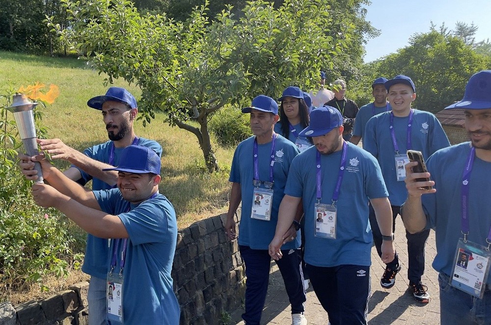 Das dritte Foto zeigt acht Athlet*innen aus der irakischen Delegation beim Fackellauf unter einen strahlend blauem Himmel. Die brennende Fackel wird von zwei Athleten getragen. 